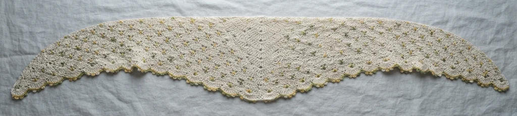 A long off-white crocheted wrap with repeating fan and picot stitch motifs laid flat on pale linen fabric, showing small yellow and green flecks in the yarn and a scalloped lower edge.