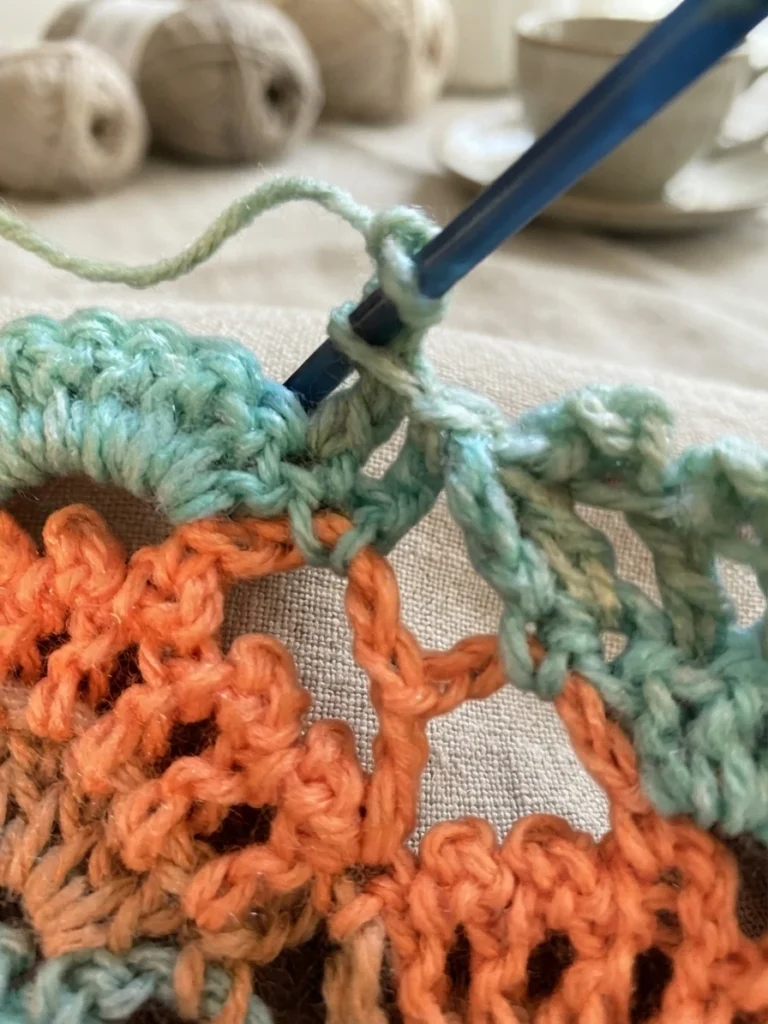 Close-up of a crochet picot in progress: a blue crochet hook is inserted into the top of a mint green double crochet to close the picot, above rows of mint green and orange lace stitches, with blurred balls of yarn and a cup in the backgrou