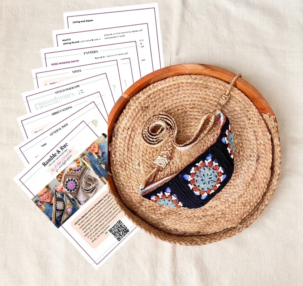 A finished crochet crossbody bag with colorful granny square motifs resting on a round woven tray, with the printed pattern pages fanned out beside it.