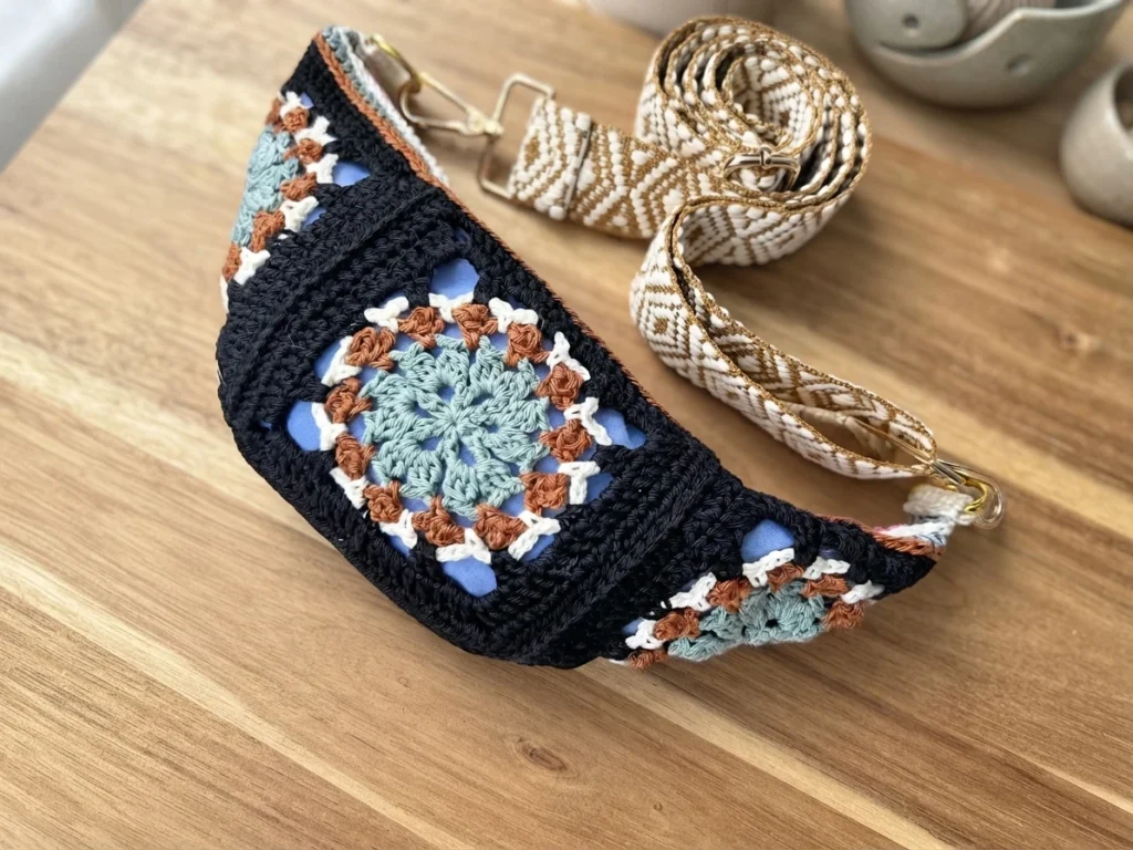 Crocheted crossbody bum bag with colorful granny square motifs in black, teal, rust and white, a light blue lining visible through the openwork, and a woven beige strap resting on a wooden table.