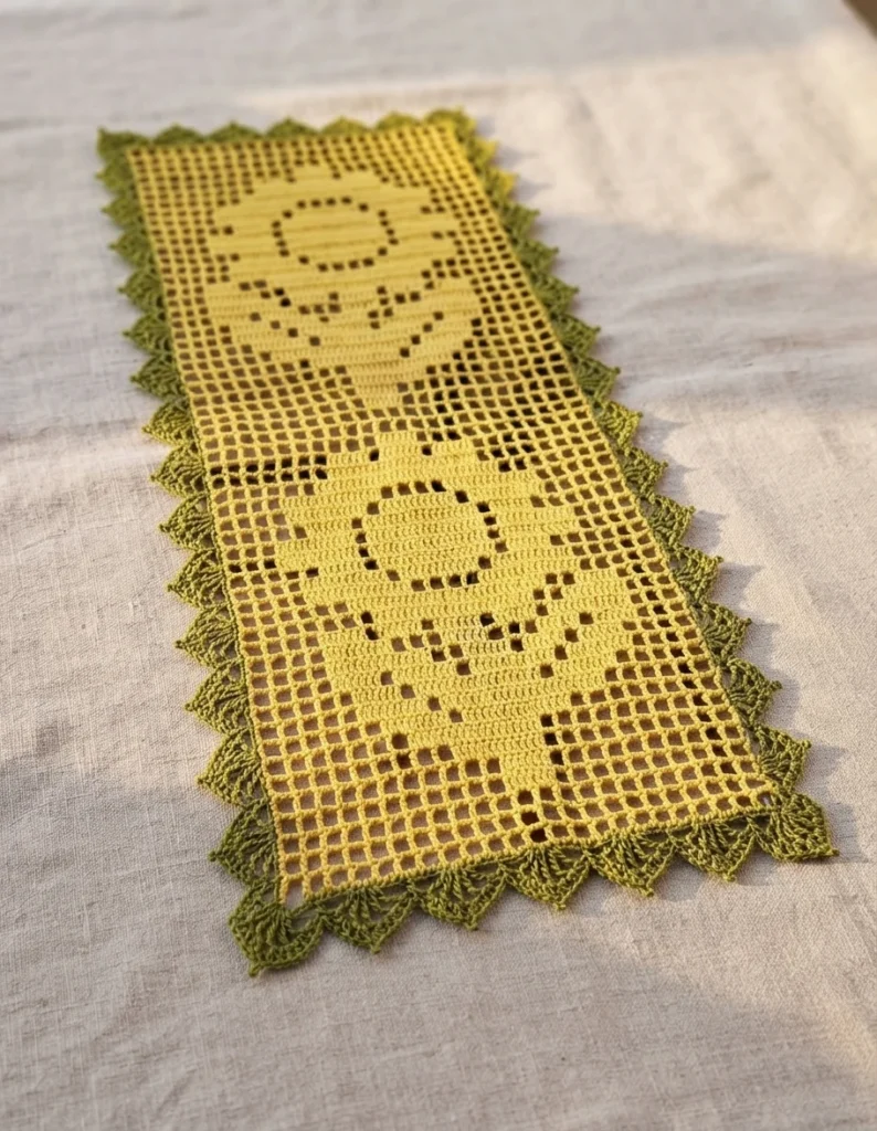 Yellow filet crochet table runner with two sunflower motifs worked in open and solid mesh blocks, edged with a green scalloped border, laid on a light linen surface.