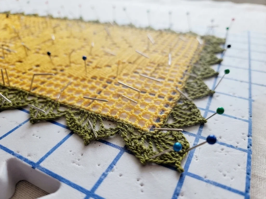 Yellow filet crochet table runner with a green scalloped edging pinned out flat on a blue grid blocking mat