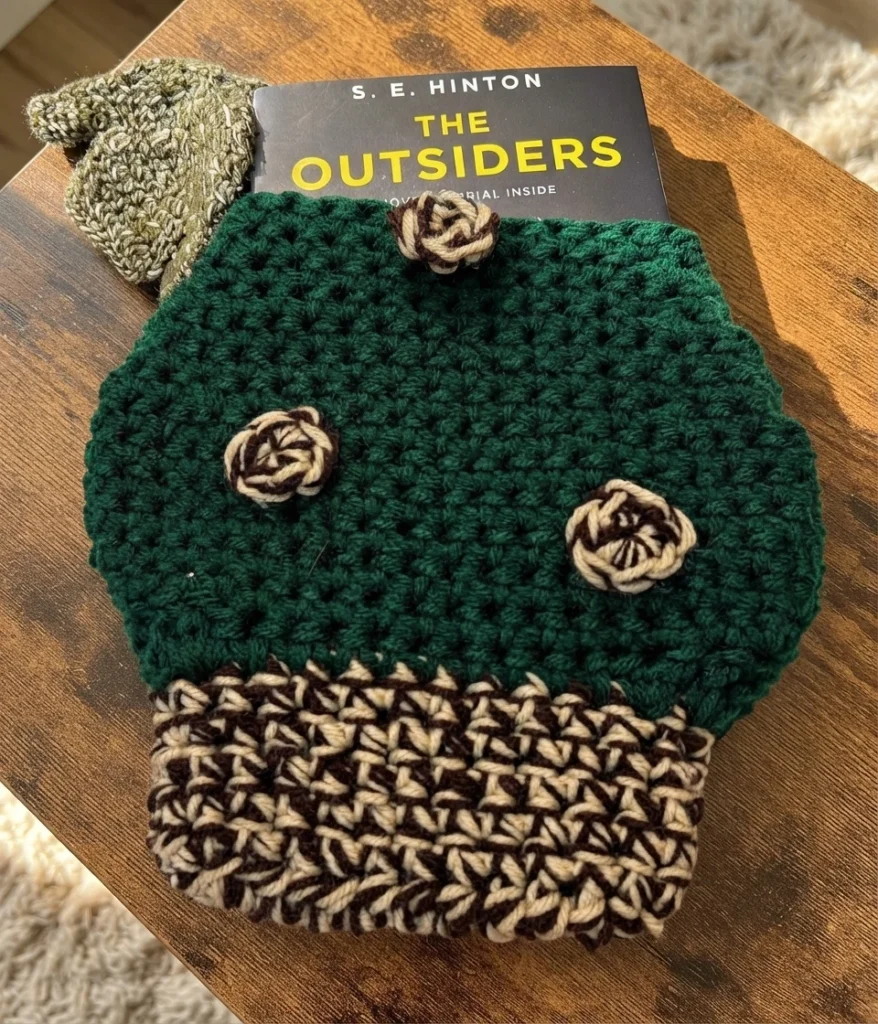 Dark green crochet mushroom book sleeve with a brown and cream mottled base and rosette spots, holding a copy of The Outsiders on a wooden table