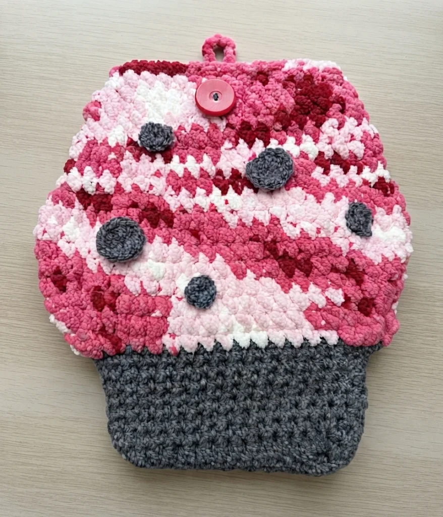 Crochet mushroom book sleeve with a pink, red and white cap, grey spots, a red button closure and a grey stem base on a light wood surface