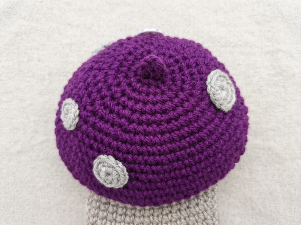 Close-up top view of a purple crochet mushroom cap with light gray spots and a beige stem on a light fabric background
