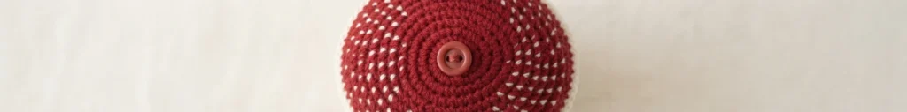 Close-up top view of a red crocheted mushroom cap with white flecks and a central pink button on a light background