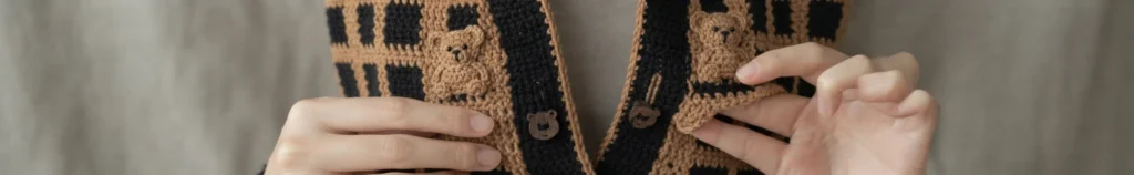 Close-up of two hands holding the front band of a crocheted cardigan in black and caramel plaid, with two small crocheted bear appliques and wooden bear-shaped buttons along the button band.