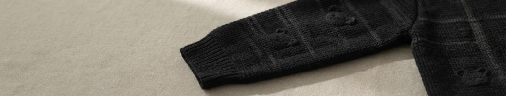 Black crocheted sleeve piece with a checkered grid texture and small raised bear motifs, laid flat on a light beige fabric surface.