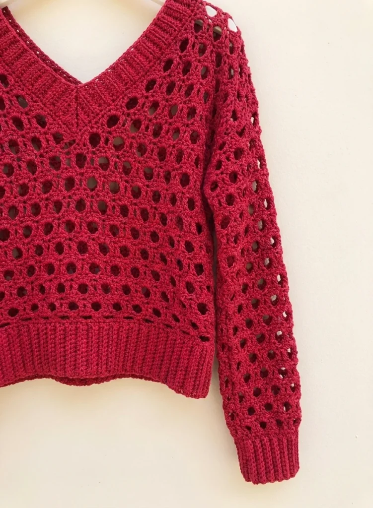 Red crochet V-neck sweater with openwork pattern and ribbed hem hanging against a light wall