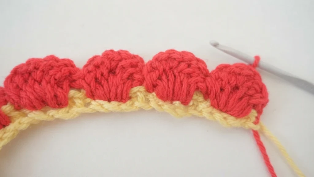 Close-up of a crocheted strip with a pale yellow chain base and coral red scalloped petals, with a light gray crochet hook and loose red and yellow yarn ends on a white background.