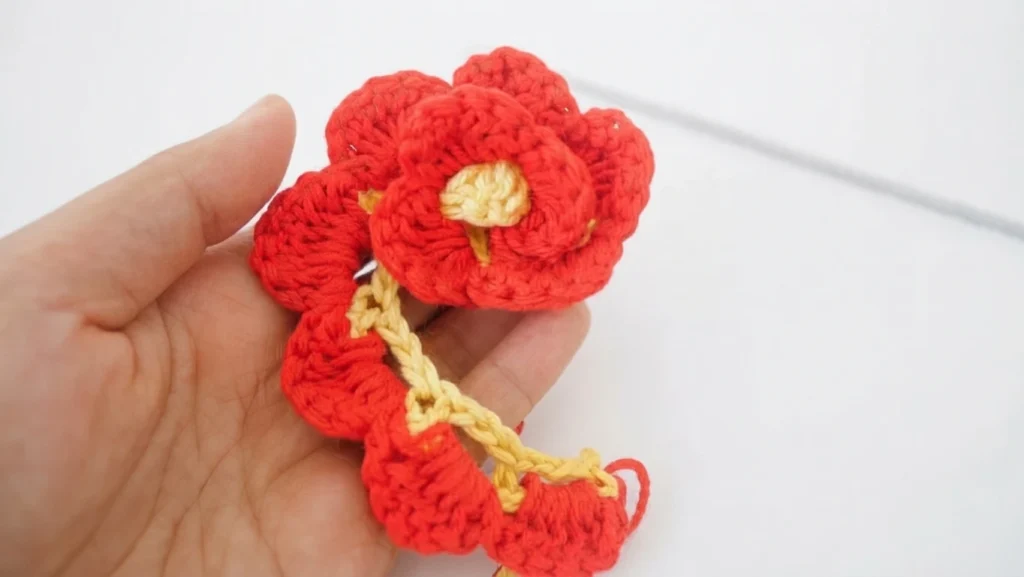 Hand holding a red crochet flower with a yellow center and a trailing petal strip.