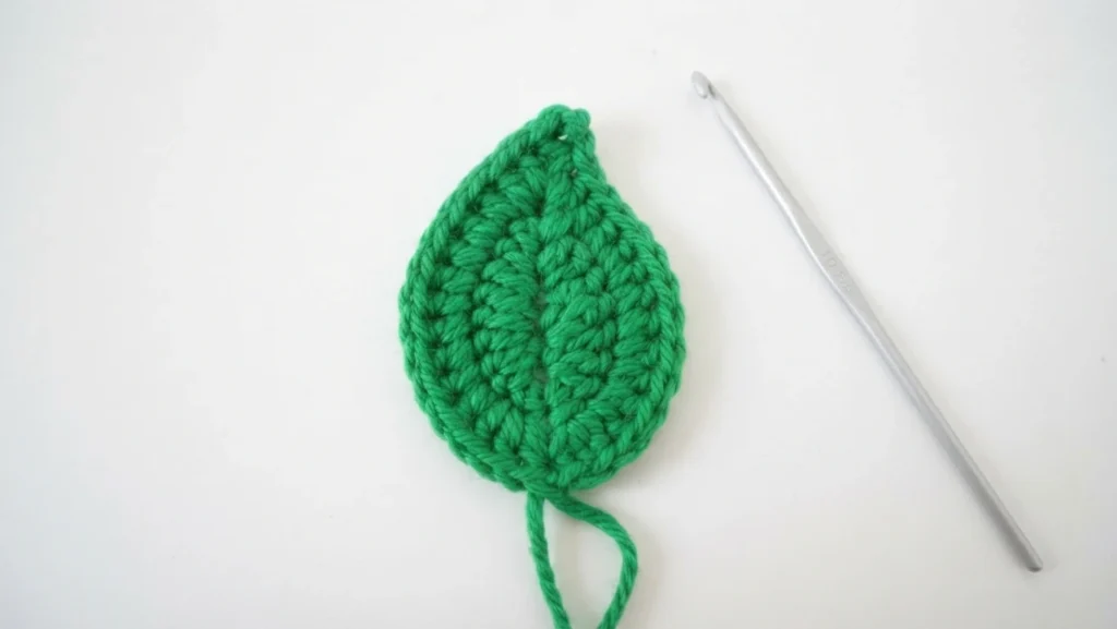 Green crocheted leaf next to a silver crochet hook on a white background.