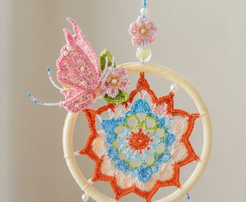 Crocheted pink butterfly with pearl beads resting on a yarn-wrapped hoop that frames an orange, blue and cream star mandala, with a beaded pink flower hanging above