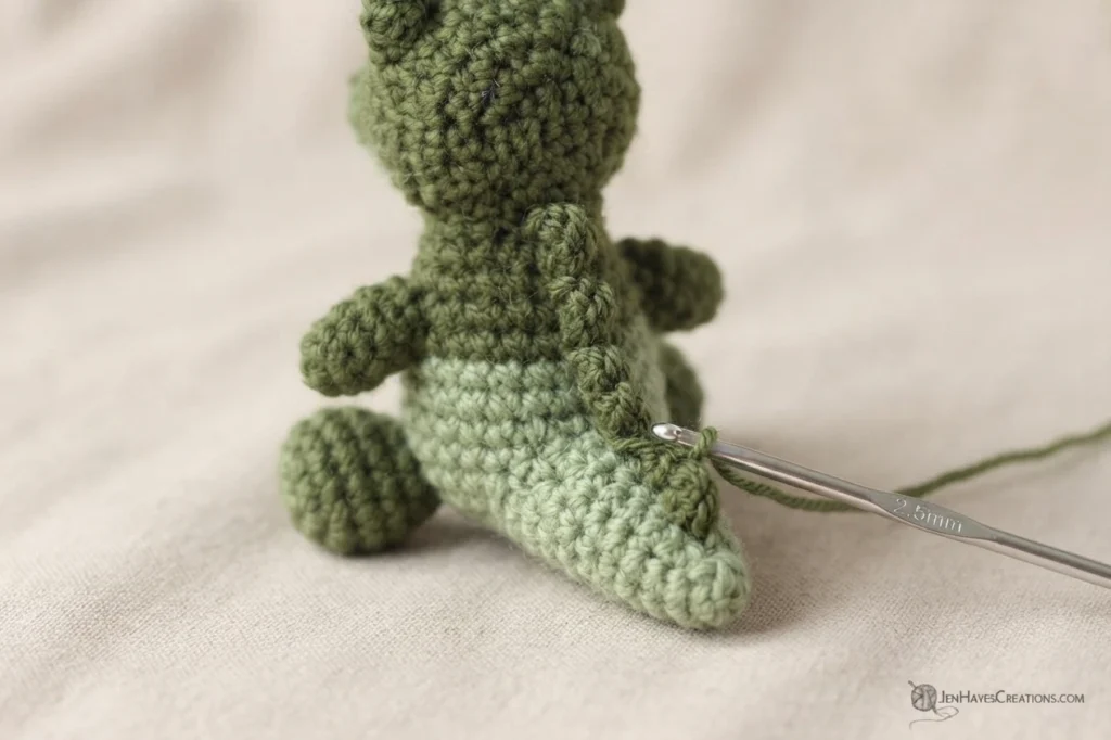 Back view of a small crocheted alligator in progress, with a metal crochet hook inserted at the tail where a row of spikes is being worked.