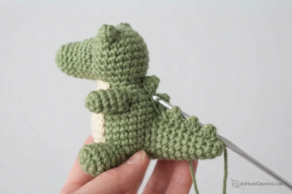 Hand holding a small green crochet alligator with a cream belly while a metal crochet hook forms small spikes along its tail and back.