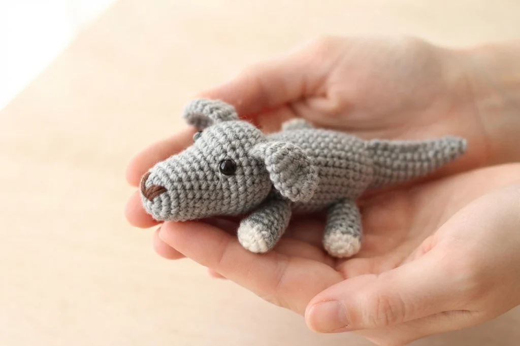 Small grey crochet animal with brown nose and white-tipped feet resting in two cupped hands.
