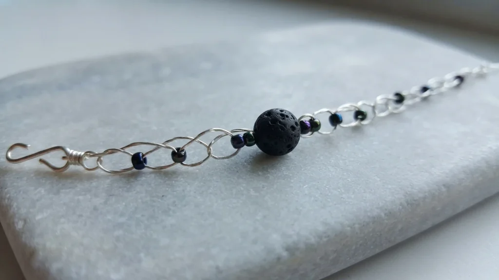 Wire crochet bracelet with one black lava rock bead, small iridescent seed beads and a wire hook clasp, lying on a light gray stone.
