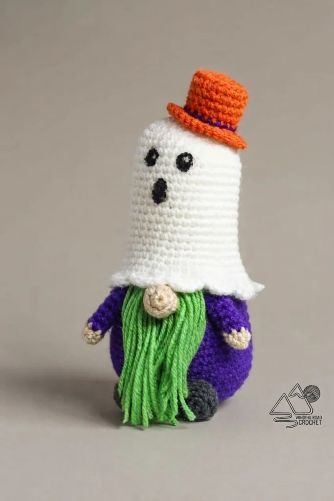 Crocheted gnome with a white ghost-shaped hat featuring embroidered black eyes and mouth, topped by an orange top hat with a purple band, green yarn beard, beige nose, purple body, beige hands and dark grey feet, photographed on a plain gre