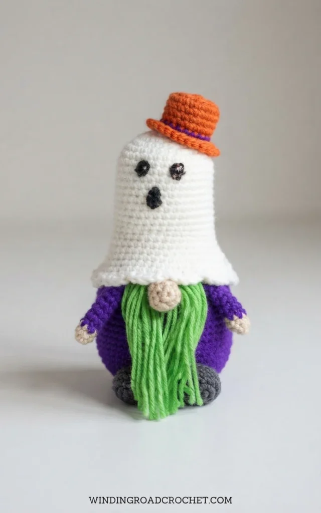 Crochet ghost gnome with a white ghost-shaped hat, a small orange top hat with a purple band, a purple body, a green yarn beard, a beige nose, and dark feet, photographed on a plain light background with a text watermark at the bottom.