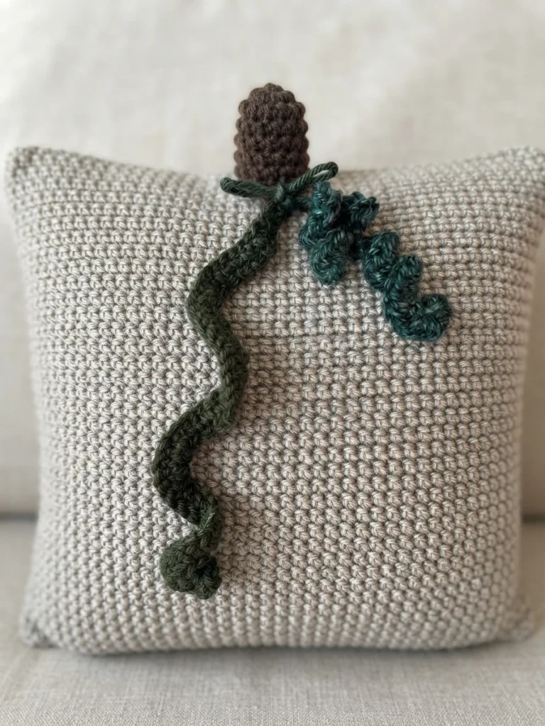 Light gray crochet pillow with a brown crocheted stem at the top and green crocheted vines trailing across the front, photographed on a pale sofa.