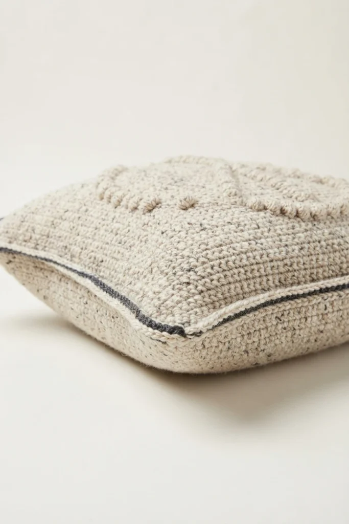 A cream crocheted pillow with a raised bobble motif, photographed at an angle on a plain light background, showing a dark gray contrast trim along the seamed edge.
