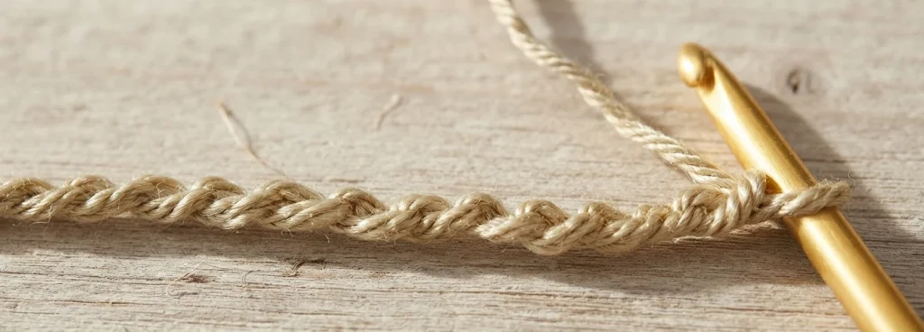 Close-up of a light beige crocheted chain on a pale wood surface, with a gold crochet hook inserted into the last chain and the working yarn trailing upward