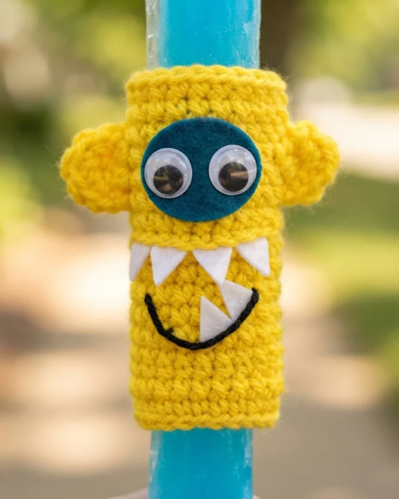 Close-up of a yellow crochet monster ice pop holder with two googly eyes and white felt teeth on a blue ice pop