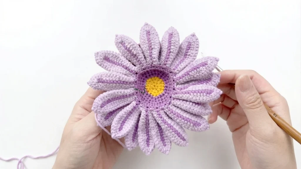 Hands holding the crocheted daisy from the back, showing light purple medium petals with darker purple center stripes around a yellow and purple center, with a wooden-handled crochet hook alongside.