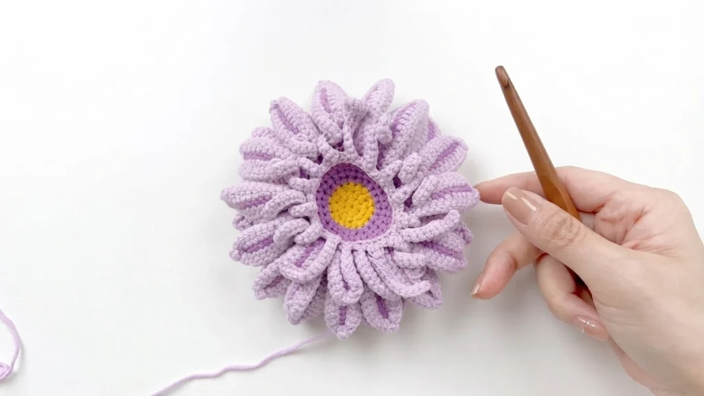 Close-up of a lavender crocheted gerbera daisy in progress, a ring of medium petals worked around a purple and yellow center, with a hand holding a wooden crochet hook beside it.