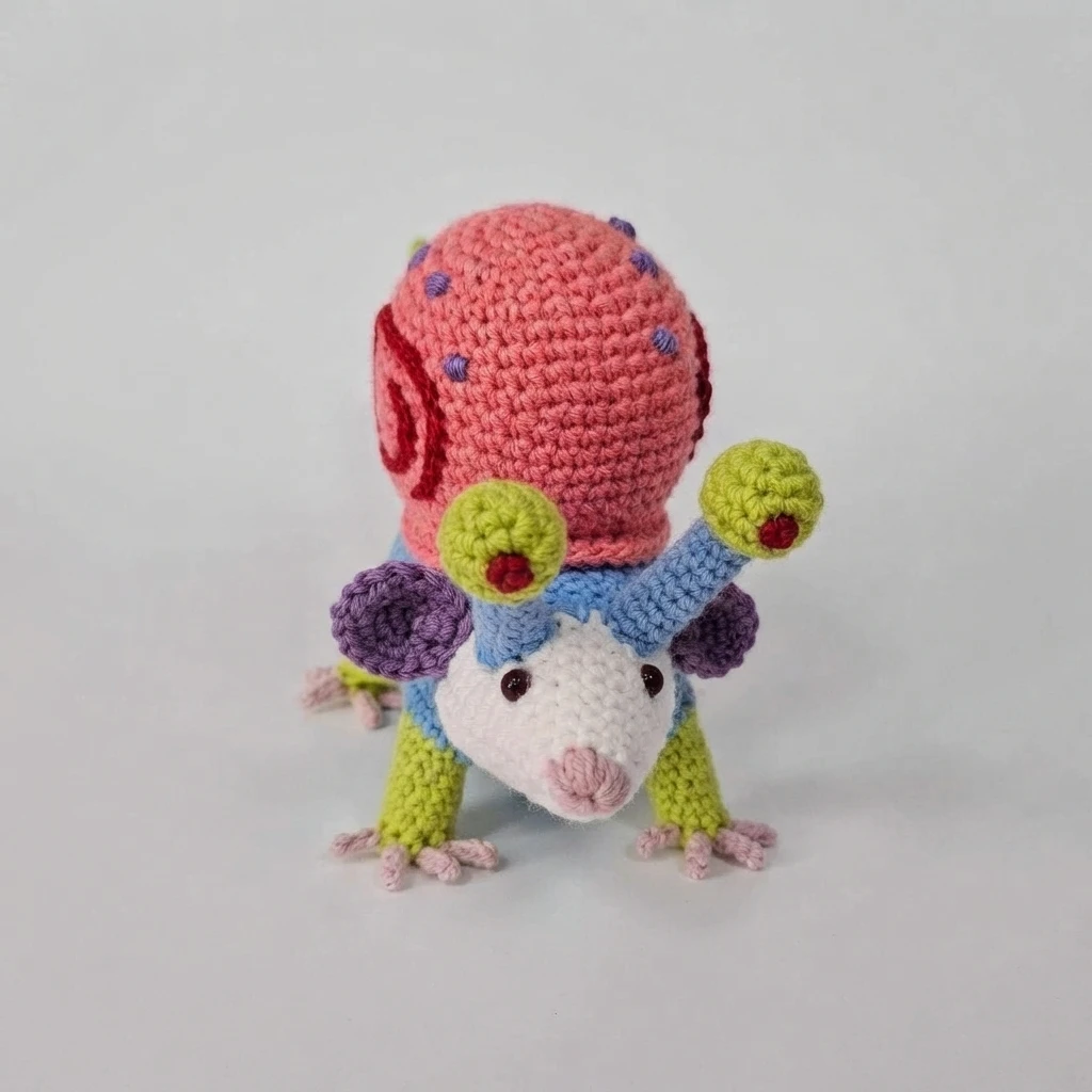 Finished crocheted possum with a white face, blue body, green limbs and eye stalks, and a pink shell decorated with purple spots.