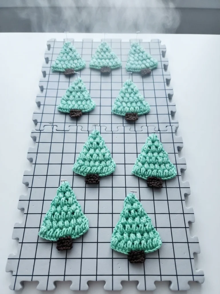 Nine crocheted green Christmas trees with dark brown trunks pinned to grey grid blocking mats, with steam rising above them