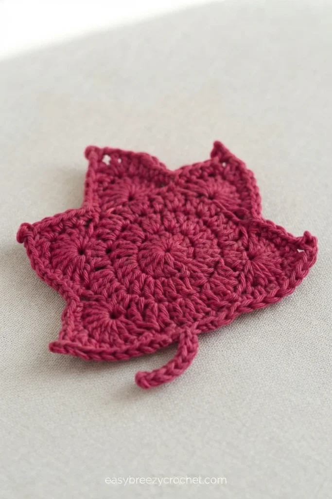 Single dark pink crocheted leaf coaster with pointed maple-style edges and a short yarn tail, resting on a plain light gray fabric surface with a small website watermark at the bottom.