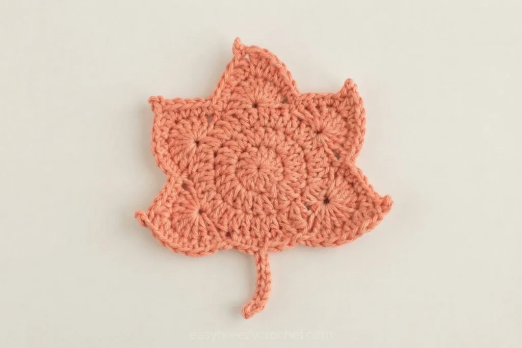 Single crochet maple leaf coaster in peach yarn with a small stem, worked in double crochet rounds forming five lobes, photographed flat on a plain light background.