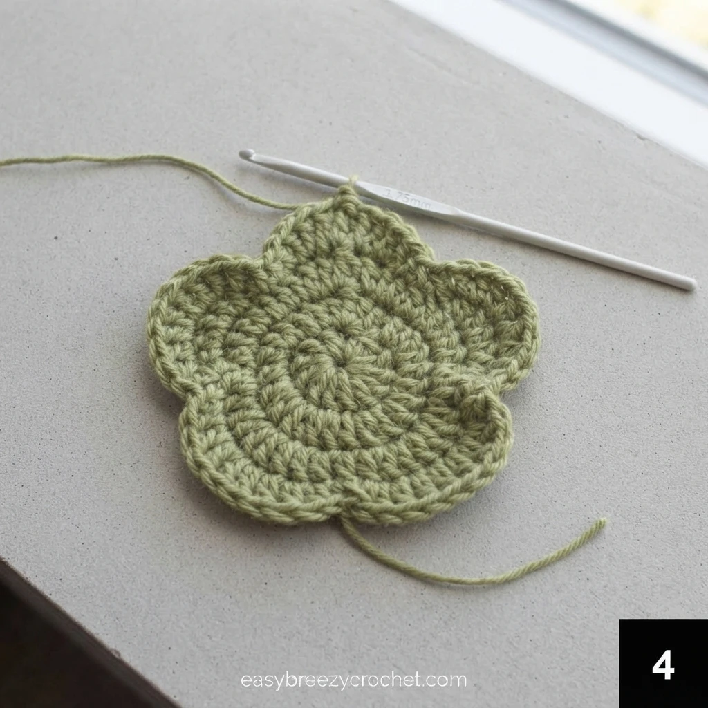 Green crocheted piece with rounded petal-like lobes and a white crochet hook resting on a pale gray surface beside a window, with loose yarn tails at the top and bottom.