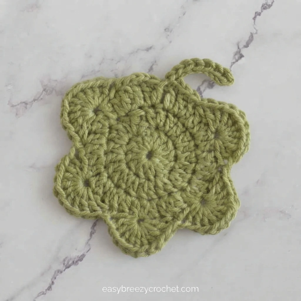 Green crochet maple leaf coaster with five rounded lobes and a short curved crocheted stem, lying flat on a white marble surface with grey veining.