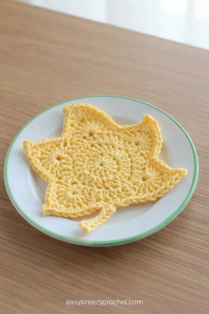 A yellow crocheted maple leaf coaster resting on a white plate with a green rim, placed on a light wooden table, with a small website watermark near the bottom.