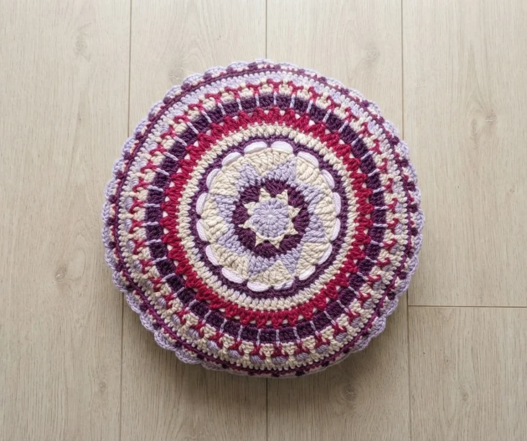 Round crochet mandala pillow in purple, pink, cream and lavender with concentric rings, viewed from above on a light wooden floor.