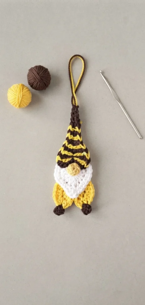 Crochet bee gnome ornament with a yellow and brown striped hat, white beard, yellow body and brown feet, hanging from a loop next to two yarn balls and a metal crochet hook on a beige surface.