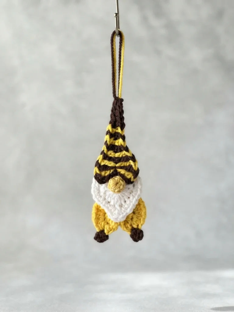 Crocheted bee gnome ornament hanging from a metal hook, with a brown and yellow striped pointed cap, a white heart shaped beard, a small light beige nose, and a yellow body with brown feet.