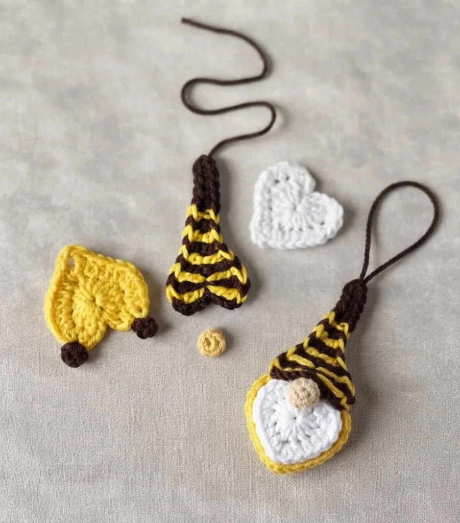 Crocheted bee gnome pieces laid on a light gray surface: a yellow body with two dark brown feet, a brown and yellow striped hat with a long brown yarn tail, a white heart shaped beard, a small cream nose, and one assembled gnome ornament wi