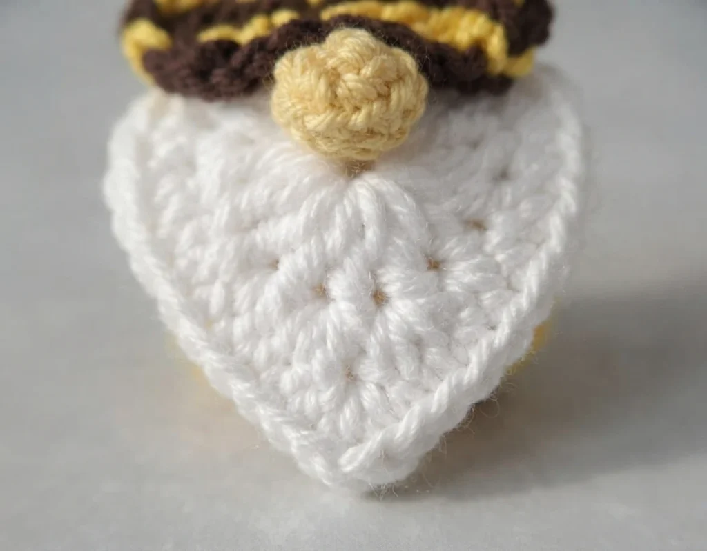 Close-up of a crocheted gnome's white heart-shaped beard with a small pale yellow nose, below the brown and yellow striped brim of a pointed hat.