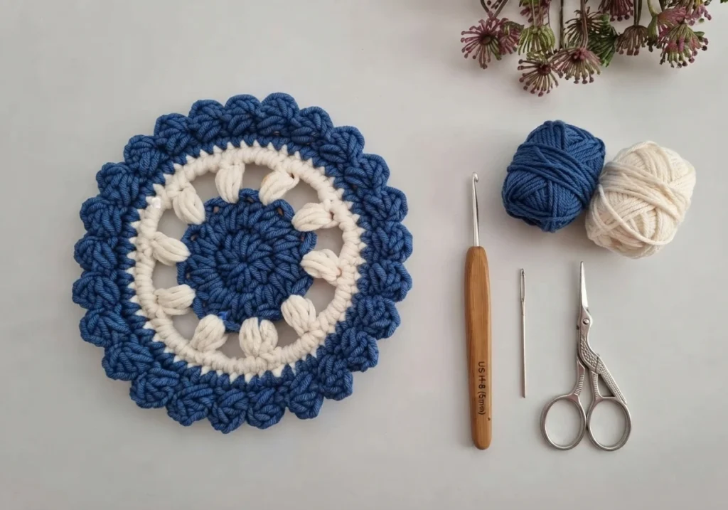 A single blue and cream round crochet coaster beside blue and cream yarn balls, a crochet hook, a tapestry needle and scissors on a pale surface