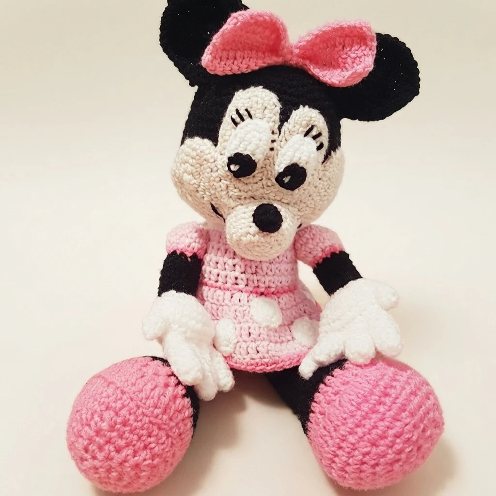 Crocheted Minnie Mouse plush with black ears, pink bow, pink polka dot dress and pink shoes, seated facing forward.