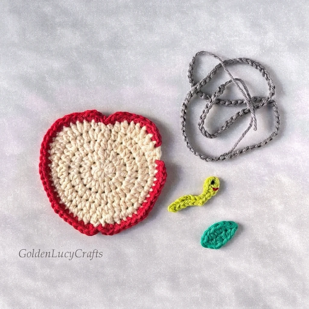 Disassembled crochet apple applique pieces on a light textured surface: a cream and red apple slice, a coiled gray chain for the worm tunnel, a small lime green worm with a red mouth, and a teal leaf.