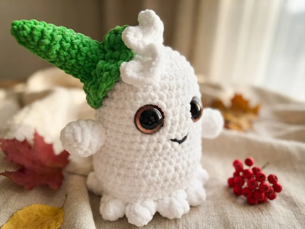 Fantôme amigurumi blanc au chapeau pointu vert, avec de grands yeux foncés et un petit sourire, posé parmi des feuilles et des baies rouges.