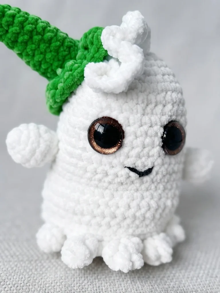 Fantôme blanc au crochet avec de grands yeux, un sourire brodé et un chapeau vert, posé sur un tissu clair.