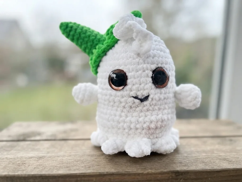 A white crocheted ghost with large dark eyes, a stitched smile, outstretched arms, and a bright green pointed hat, standing on a wooden surface.