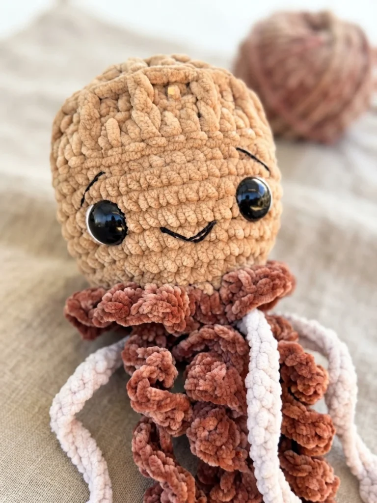 A brown crocheted jellyfish with a smiling face, curled tentacles and long white tentacles, photographed on a light surface.