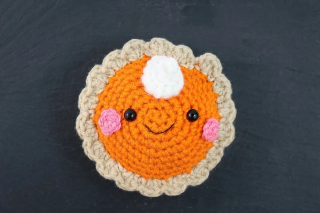 Crocheted pumpkin pie amigurumi with an orange filling, a beige scalloped crust, a white whipped cream swirl on top, pink cheeks, black safety eyes and an embroidered smile, photographed on a dark surface.