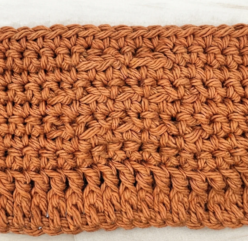Close-up of a flat crocheted panel in orange cotton yarn, showing dense rows of single crochet stitches with raised front post stitches forming a textured pattern, photographed on a light fabric background.